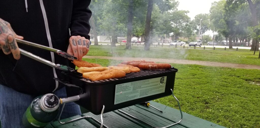 Cooking sausages on a small tabletop grill at a park picnic table
